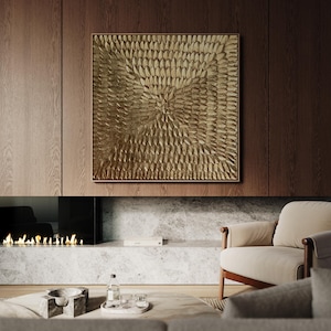 A large square abstract canvas painting featuring a heavy 3D textured gold leaf surface with a radiating sunburst pattern. Displayed in a bright, luxury modern living room above a classic white fireplace.