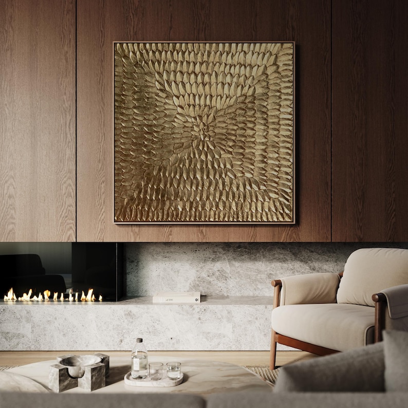 A large square abstract canvas painting featuring a heavy 3D textured gold leaf surface with a radiating sunburst pattern. Displayed in a bright, luxury modern living room above a classic white fireplace.