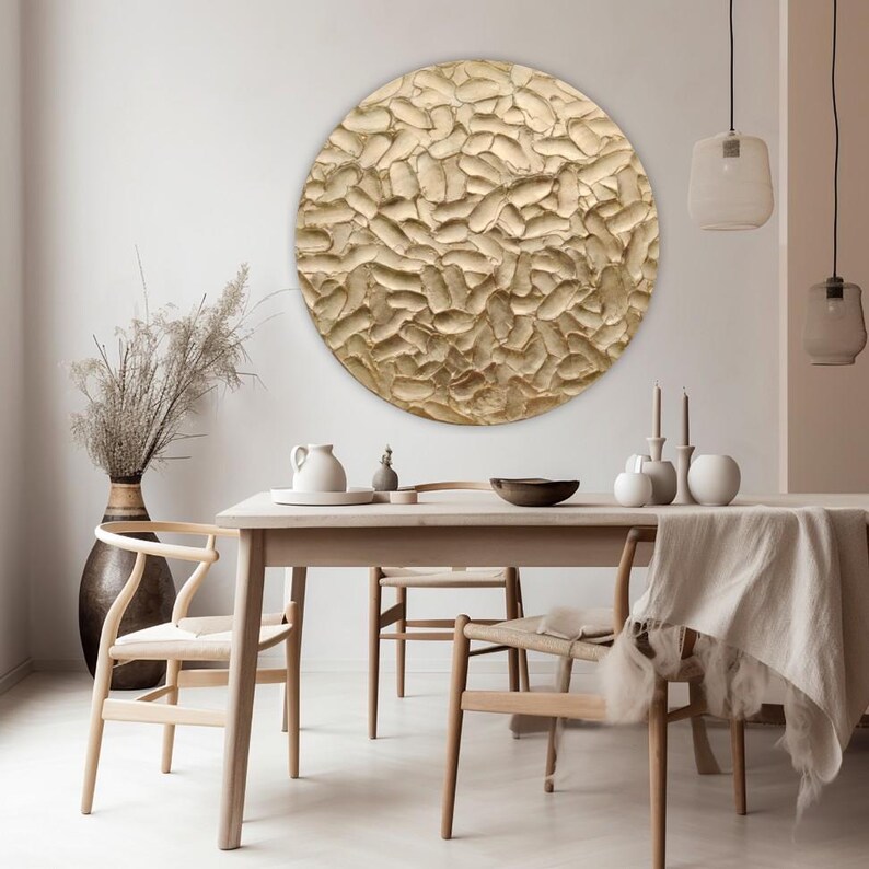 A large, round, textured wall art piece with a golden-beige color. The artwork is the focal point of a dining room, hanging above a wooden table and chairs. The room has a neutral color palette.