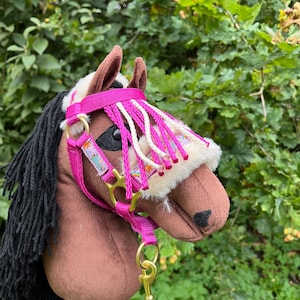 May include: A brown hobby horse with a black mane and a pink bridle. The bridle features a decorative design and is accented with white and pink fringe. The horse has a soft, plush appearance and is designed for play.