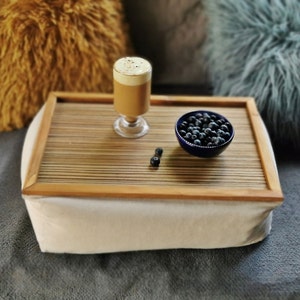 Puede incluir: Una bandeja de madera con una base de cojín blanco, un vaso de café y un pequeño cuenco azul lleno de arándanos. La bandeja está sobre una superficie gris y peluda.