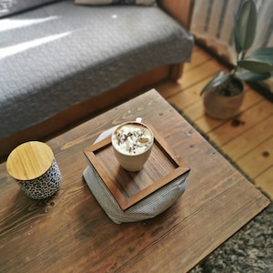 Può includere: Un vassoio di legno con una tazza di caffè bianca con panna montata e granella di cioccolato sopra, appoggiato su un cuscino di stoffa a righe. Il vassoio è su un tavolo di legno con un barattolo di legno con coperchio.