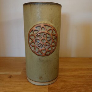 Könnte beinhalten: Eine zylindrische, olivgrüne Keramikvase mit einem erhabenen, kreisförmigen, kupferfarbenen Blumendesign. Die Vase hat eine leicht strukturierte Oberfläche und einen dunkelbraunen Rand. Die Vase steht auf einer Holzoberfläche.