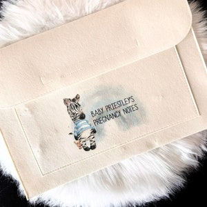 May include: A white felt folder with a zebra illustration and the text "Baby Priestley's Pregnancy Notes".