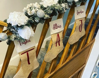 May include: Four personalised burlap stockings with red lettering hanging on a wooden banister decorated with white and green garland. The stockings are labelled "Tasha", "Albie", "Jordan", and "J".