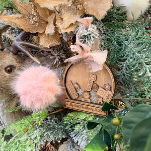 Peut inclure: Ornement de Noël en bois avec le texte "Name" et "My First Christmas 2023". L'ornement rond présente des motifs découpés d'objets pour bébés. Il est décoré d'un nœud rose et d'un pompon rose.