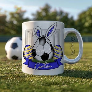May include: White ceramic mug featuring a football wearing bunny ears and surrounded by Easter eggs. The mug has a blue ribbon with the name "Carter" on it. The mug is on a green grassy surface.