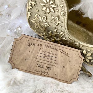 Peut inclure: Un billet officiel du Père Noël en bois avec le nom "DARCIE" et le texte "NICE LIST" est affiché. Le billet est posé sur une surface blanche et duveteuse, avec un traîneau doré en arrière-plan. Le billet mesure environ 15 cm de long.