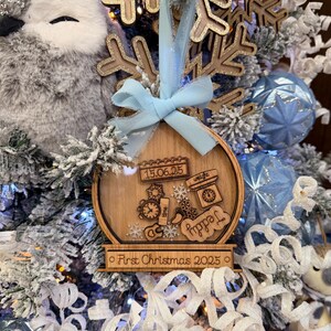 Peut inclure: Ornement de Noël en bois avec un centre transparent, avec des détails gravés comme une horloge, des flocons de neige et le mot "Teddy". L'ornement est orné d'un ruban bleu clair et du texte "First Christmas 2025."