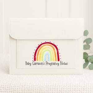 May include: White leather folder with a rainbow design and the text "Baby Garason's Pregnancy Notes".