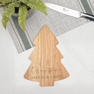 May include: A wooden, tree-shaped chopping board with the text "Off my tits on... M&S PICKY BITS" engraved on it. The board is light brown and sits on a grey and white striped placemat. A stainless steel knife is visible.