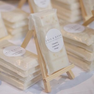 May include: A stack of white bath and body bars wrapped in clear plastic with a label that reads "Iris & Rose, Hand Baths and Essential Oils, Cinnamon, Cranberry & Orange". The bars are displayed on a small wooden easel.