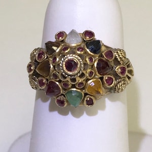 May include: A gold ring with a cluster of colorful gemstones, including red, green, blue, and yellow. The ring is set with a variety of shapes and sizes of stones, and the design is intricate and detailed.