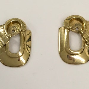 May include: A pair of gold-toned earrings with a unique geometric design. The earrings are shaped like a U with a smaller, curved shape attached to the top.