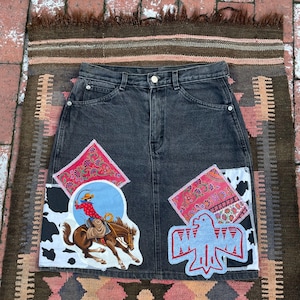 May include: A black denim skirt with colourful patches. The patches include a cowboy riding a horse, a bird, and a paisley pattern.