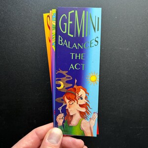 May include: A bookmark with a colorful illustration of a woman with two faces, one with a sun and the other with a moon. The text "GEMINI BALANCES THE ACT" is printed on the bookmark.