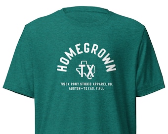 May include: A teal t-shirt with the text "HOMEGROWN TX" in a white arched font. Below the text is "TRICK PONY STUDIO APPAREL CO. AUSTIN TEXAS, Y'ALL" in a white font. The shirt is made of a soft material.