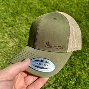 May include: Olive green Snapback hat with a tan mesh back. The hat features a brown leather patch with a mountain range design. The hat has the text "THE AUTHENTIC YUPOONG SINCE 1974" on a silver sticker.