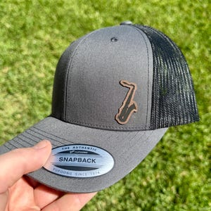 May include: Gray snapback hat with a black mesh back and a brown saxophone patch. The hat has a curved brim and a silver tag that reads "The Authentic Snapback Yupoong Since 1974".