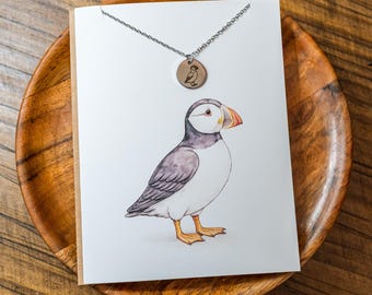 May include: A silver-toned chain necklace with a small circular pendant featuring a puffin illustration, displayed on a white card printed with a detailed watercolor-style puffin bird with black, white, and orange markings.