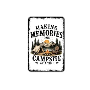 May include: A rectangular metal sign with a white background and distressed black edges. The text reads "MAKING MEMORIES ONE CAMPSITE AT A TIME" in black. The illustration features a tent, a campfire, a wooden chair, pine trees, and mountains.