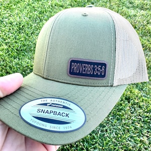 May include: Olive green snapback hat with a tan mesh back. The hat features a rectangular patch with the text "PROVERBS 3:5-6". The brim has a sticker that reads "THE AUTHENTIC SNAPBACK YUPOONG SINCE 1974".