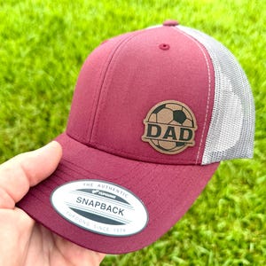 May include: Maroon and gray trucker hat with a soccer ball and the word "DAD" on a leather patch. The hat has a curved brim and a mesh back. The tag reads "The Authentic Yupoong Snapback Since 1974".