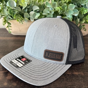 May include: A gray trucker hat with a black mesh back. The hat features a brown leather patch with the text "IN JESUS NAME". The brim is gray with a black stitched design. A black and red label is on the brim.