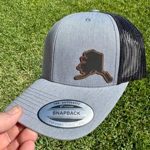 May include: Gray snapback hat with a black mesh back and a brown leather patch in the shape of Alaska. The hat has a curved brim and a metal tag that reads "SNAPBACK" and "YUPOONG SINCE 1974".