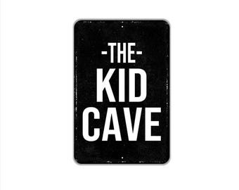 May include: A rectangular black metal sign with rounded corners featuring the white text -THE- KID CAVE in a bold sans-serif font. The surface has a subtle distressed texture with small mounting holes visible at the top and bottom center.