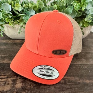 May include: A coral-colored Snapback baseball cap with a brown patch and a tan mesh back. The cap features a curved bill and a white and black tag that reads "THE AUTHENTIC SNAPBACK YUPOONG SINCE 1974."