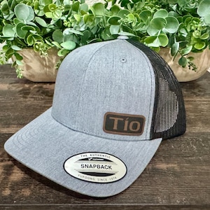 May include: A gray Snapback baseball cap with a black mesh back. The cap features a brown leather patch with the word "Tío" and a white and black patch that reads "The Authentic Snapback Yupoong Since 1974".