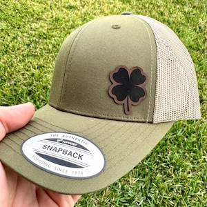 May include: Olive green snapback hat with a tan mesh back and a four-leaf clover patch. The hat features the text "SNAPBACK" on a white and black oval label. The hat is made by Yupoong since 1974.