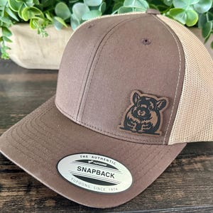 May include: Brown snapback hat with a tan mesh back and a patch featuring a black and brown animal illustration. The hat has a curved brim and a tag that reads "The Authentic Snapback Yupoong Since 1974."