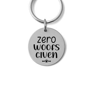 May include: A round, silver-colored dog tag with a key ring. The tag has the phrase "zero woofs given" in black, bold lettering. Below the text is a paw print and bone graphic.