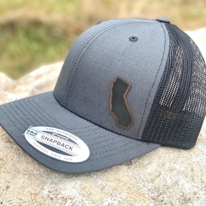 May include: A gray snapback trucker hat with a black mesh back. The hat features a brown leather patch in the shape of California. The hat has a curved brim and a white and black tag that reads "Yupoong Since 1974".