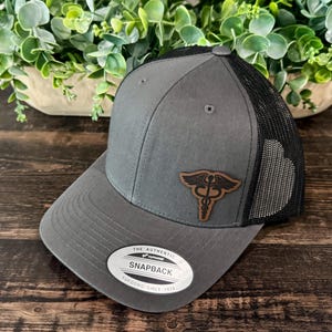May include: A gray snapback hat with a black mesh back. The hat features a brown leather patch with a medical caduceus symbol. The hat has a curved brim and a snap closure. The text "THE AUTHENTIC YUPOONG SINCE 1974" is printed on a white oval sticker.