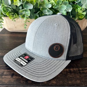 May include: Gray trucker hat with a black mesh back and a curved brim. The hat features a leather patch with an eight ball design. The tag reads "RICHARDSON 112 TRUCKER".