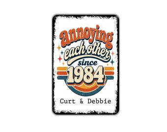 May include: A rectangular metal sign with a white background and distressed black edges. It features the text "annoying each other since 1984" in orange and blue, with "Curt & Debbie" printed in black below a colorful retro-style graphic.