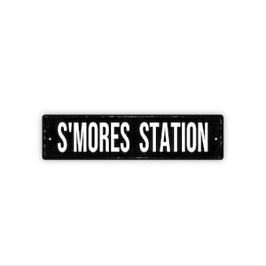 May include: A rectangular black metal sign with the words "S'MORES STATION" in white, centered on the sign. The sign is set against a white background.
