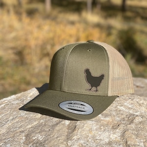 May include: Olive green and tan snapback hat featuring a brown chicken silhouette on the front. The hat has a curved brim and a mesh back. The text "THE AUTHENTIC SNAPBACK YUPOONG SINCE 1974" is on the front.
