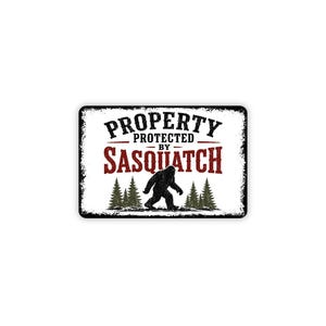 May include: A rectangular sign with a white background and a distressed black border. The text reads PROPERTY PROTECTED BY SASQUATCH in black and red lettering. A black silhouette of a Sasquatch walking among green pine trees is centered.