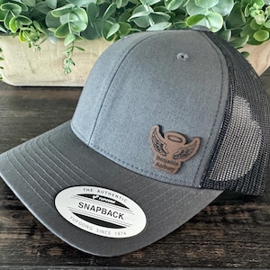 May include: A gray Snapback baseball cap with a black mesh back. The cap features a brown leather patch with an angel wing design and the name "Benjamin Anthony". The hat has a curved brim and a "Yupoong Since 1974" tag.