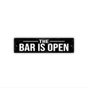 May include: A rectangular black metal sign with the words "THE BAR IS OPEN" in white, centered on the sign. The sign has a distressed, vintage look, with a white background.