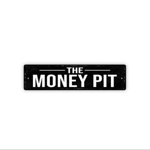May include: A rectangular black metal sign with the words "THE MONEY PIT" in white block letters. The sign has a distressed, vintage appearance, with a white horizontal line separating the two lines of text.