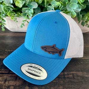 May include: A blue and white trucker hat with a fish patch. The hat features a blue front panel and brim, a white mesh back, and a brown fish emblem. The hat has a snapback closure and a tag that reads "The Authentic Snapback Yupoong Since 1974."