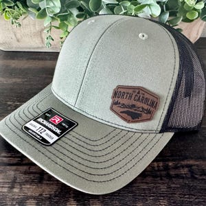May include: A green and black trucker hat with a leather patch that reads "North Carolina". The hat has a curved brim and a mesh back. The tag reads "Richardson 112 Trucker".