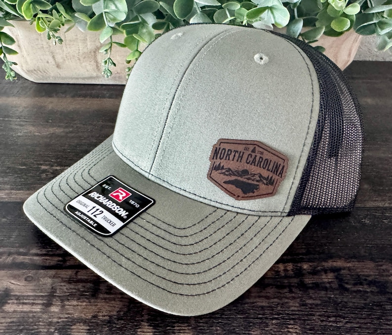 May include: A green and black trucker hat with a leather patch that reads "North Carolina". The hat has a curved brim and a mesh back. The tag reads "Richardson 112 Trucker".
