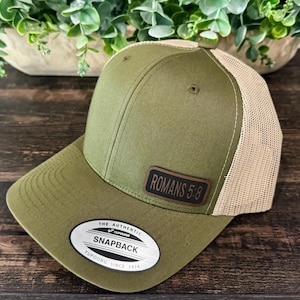 May include: Olive green Snapback baseball cap with a tan mesh back. The cap features a leather patch with the text "ROMANS 5:8". The brim is curved, and the cap has a snap closure. The tag reads "THE AUTHENTIC SNAPBACK YUPOONG SINCE 1974".