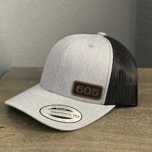 Puede incluir: Gorra snapback gris con la parte trasera de malla negra. La gorra presenta un parche de cuero marrón con el número "605" y una etiqueta "Snapback". La gorra está sobre una superficie de madera.